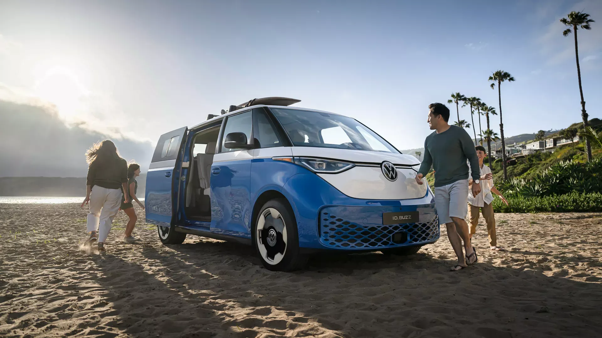 Family walking on the beach beside a blue and white 2025 Volkswagen ID. Buzz electric van near Daphne, Alabama.